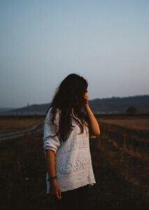 Young woman standing in a field at dusk, wearing a white knit sweater, hand raised to her face, looking away.