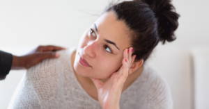Woman with dark hair in a bun rests her head on her hand while a second hand touches her shoulder, conveying support or comfort.